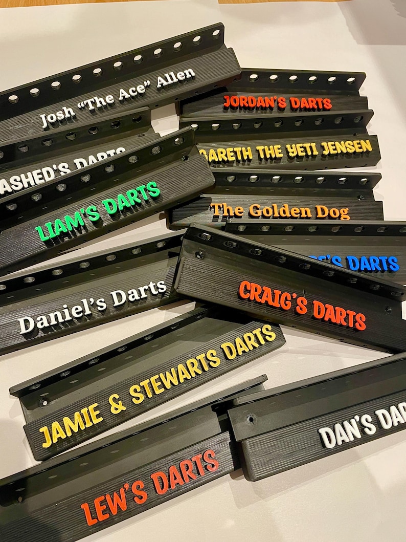 May include: A collection of black dart holders with various names in colorful 3D lettering. Names include "Josh 'The Ace' Allen", "Jordan's Darts", "Liam's Darts", "Daniel's Darts", "Craig's Darts", and others. The holders have a slotted design.