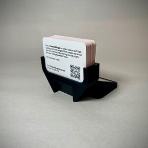 May include: A black business card holder with a stack of light pink business cards. The cards have rounded corners and text that includes a website and QR code. The holder is made of a matte black material.