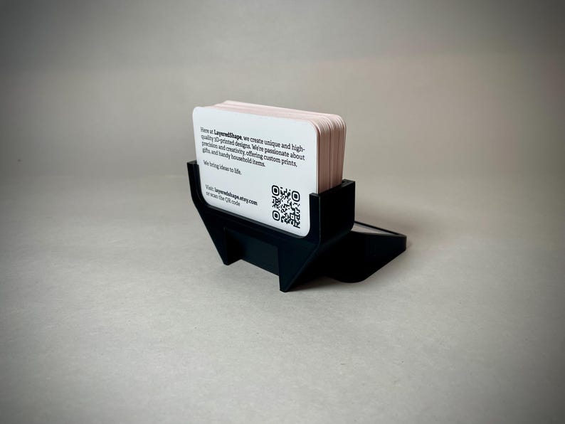 May include: A black business card holder with a stack of light pink business cards. The cards have rounded corners and text that includes a website and QR code. The holder is made of a matte black material.
