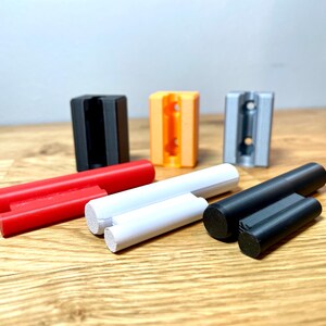 Op de afbeelding: Vier 3D-geprinte wandbevestigingen in zwart, oranje, grijs en wit, met drie verschillende gekleurde staven, rood, wit en zwart.