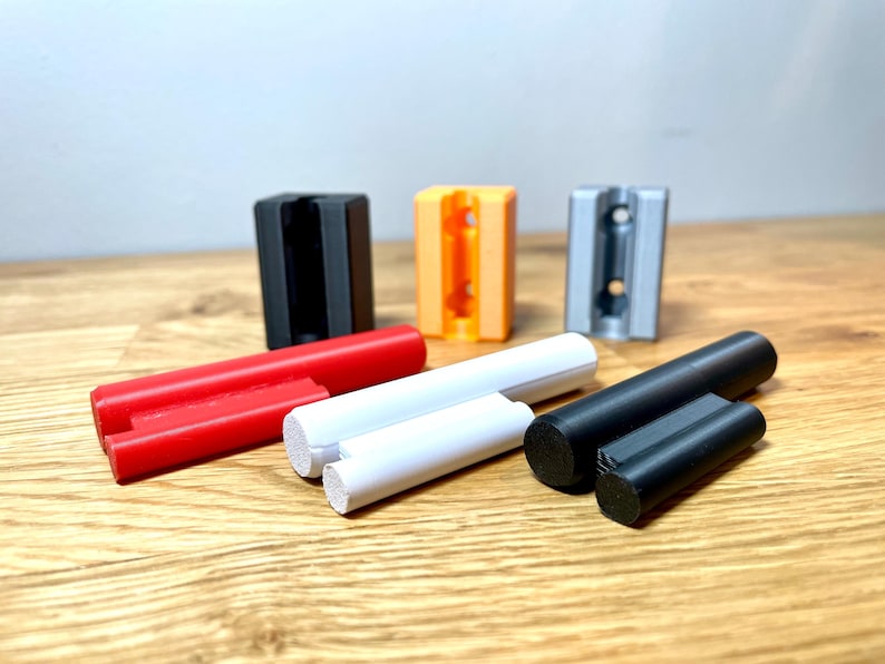 Op de afbeelding: Vier 3D-geprinte wandbevestigingen in zwart, oranje, grijs en wit, met drie verschillende gekleurde staven, rood, wit en zwart.