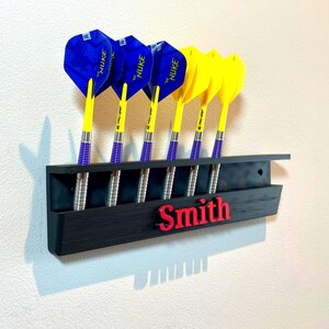 May include: A black dart holder with six darts. The darts have blue and yellow flights, silver barrels, and are labeled "NUKE". The name "Smith" is in red letters on the holder.