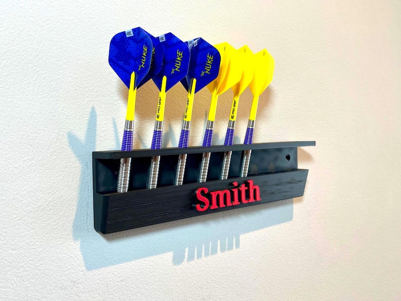 May include: A black dart holder with six darts. The darts have blue and yellow flights, silver barrels, and are labeled "NUKE". The name "Smith" is in red letters on the holder.