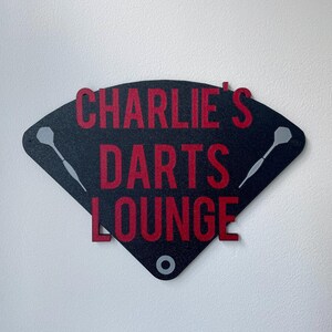 Triangular darts sign with the text charlie's darts lounge in red. Main background is black and has silver inlayed darts logos. On a white wall