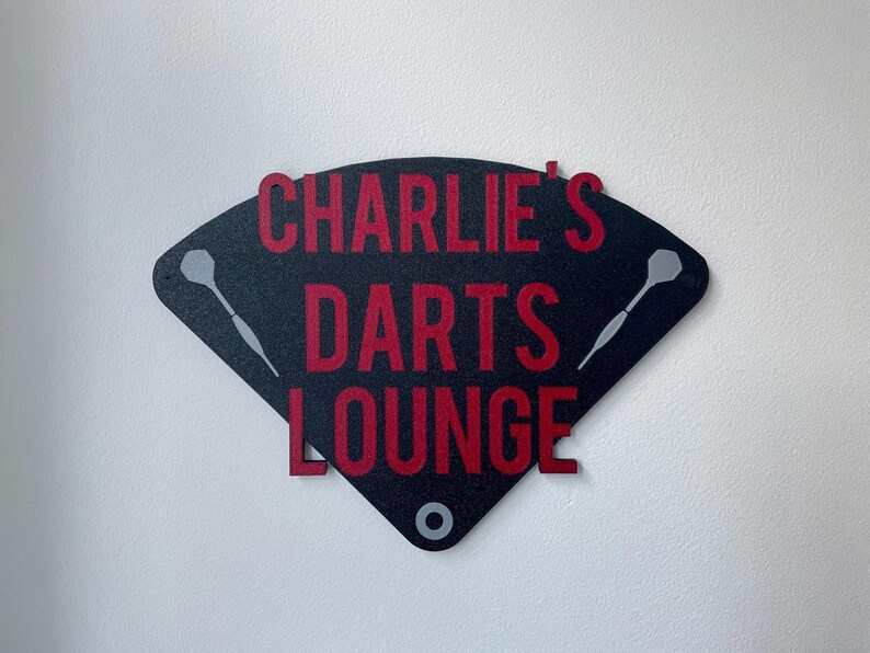 Triangular darts sign with the text charlie's darts lounge in red. Main background is black and has silver inlayed darts logos. On a white wall