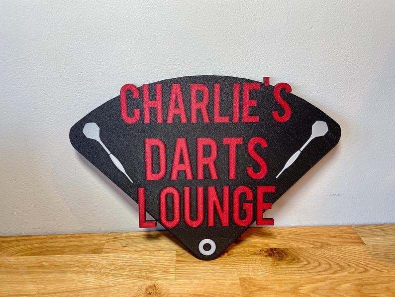 Triangular darts sign with the text charlie's darts lounge in red. Main background is black and has silver inlayed darts logos.
