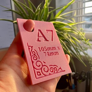 May include: A rectangular pink plastic template featuring the text A7 and 105mm x 74mm embossed on the surface. The template includes a circular hole at the top and an intricate, decorative scrollwork pattern etched into the bottom left corner.