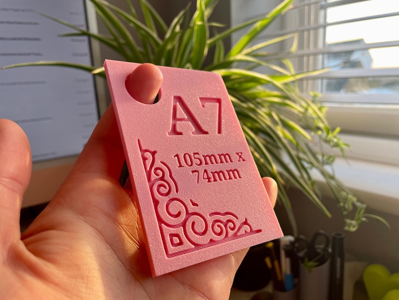 May include: A rectangular pink plastic template featuring the text A7 and 105mm x 74mm embossed on the surface. The template includes a circular hole at the top and an intricate, decorative scrollwork pattern etched into the bottom left corner.