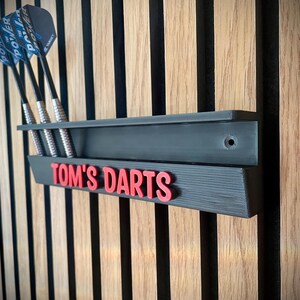 May include: A black dart holder with the text "TOM'S DARTS" in red, mounted on a wooden wall. Three darts with blue and silver details are in the holder. The holder is designed to store darts.