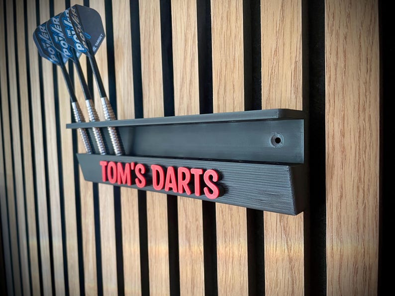 May include: A black dart holder with the text "TOM'S DARTS" in red, mounted on a wooden wall. Three darts with blue and silver details are in the holder. The holder is designed to store darts.