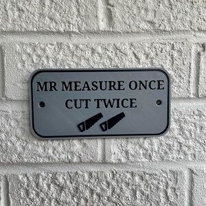 May include: A rectangular, silver-colored sign with the text "MR MEASURE ONCE CUT TWICE" in black. Two black saw icons are below the text. The sign is mounted on a white brick wall.