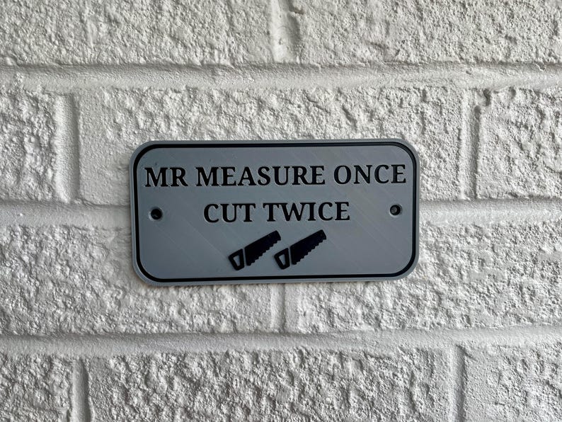 May include: A rectangular, silver-colored sign with the text "MR MEASURE ONCE CUT TWICE" in black. Two black saw icons are below the text. The sign is mounted on a white brick wall.