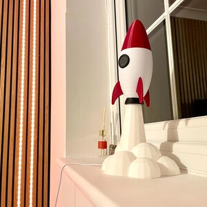 Op de afbeelding: Een witte en rode raketlamp met een zwart rond venster en rode vinnen. De lamp staat op een witte basis met wolkvormige details. Op een witte vensterbank.