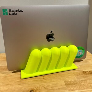 May include: A neon yellow 3D-printed laptop stand featuring five rounded, slanted vertical supports on a rectangular base, holding a silver laptop with a Bambu Lab logo and a small circular green character sticker on the back.