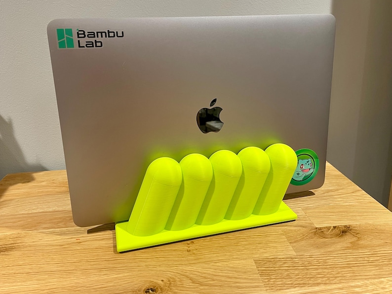 May include: A neon yellow 3D-printed laptop stand featuring five rounded, slanted vertical supports on a rectangular base, holding a silver laptop with a Bambu Lab logo and a small circular green character sticker on the back.