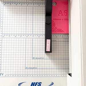 May include: A white paper cutter with a black handle labeled TOOL BOX, featuring a grid-marked base with measurements in inches and centimeters. A bright pink A6 card measuring 148mm x 105mm with decorative corner patterns is positioned under the blade.