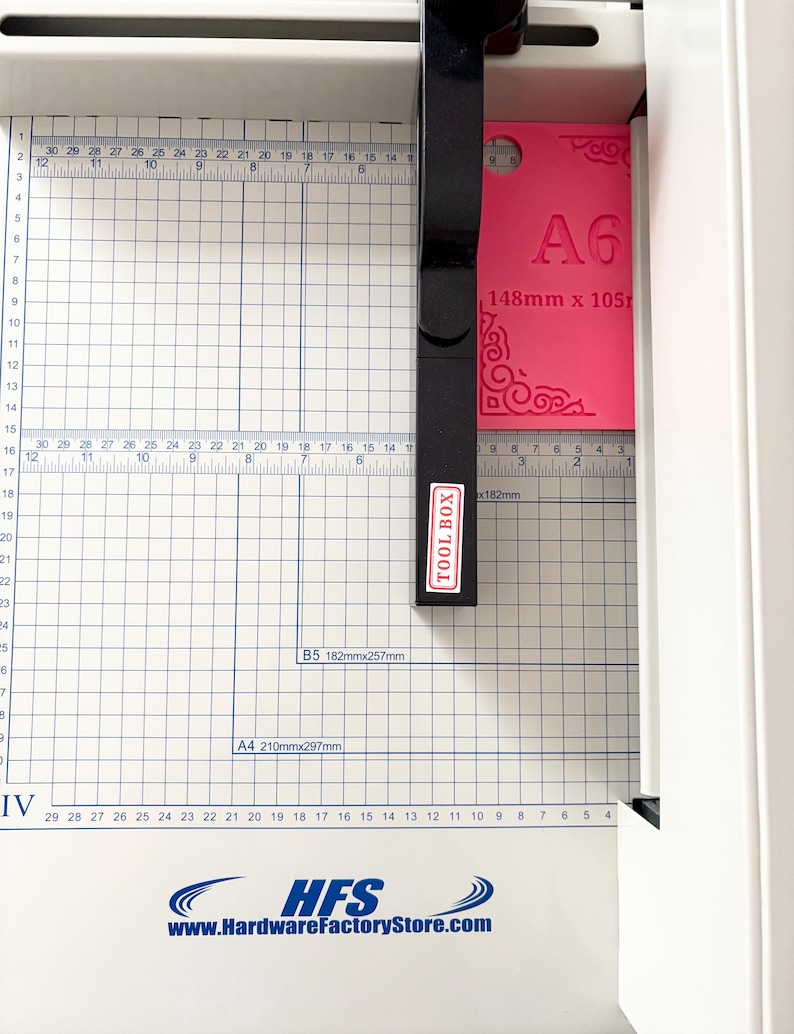 May include: A white paper cutter with a black handle labeled TOOL BOX, featuring a grid-marked base with measurements in inches and centimeters. A bright pink A6 card measuring 148mm x 105mm with decorative corner patterns is positioned under the blade.