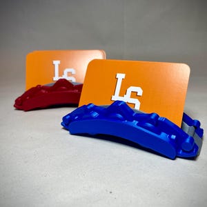 May include: Two business card holders with a car brake caliper design. One is red, the other is blue. Each holds an orange card with the white letters "Ls". The background is a neutral gray.