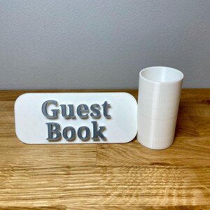 pen pot on the right with a sign that says guest book. Shown on a wooden desk with grey background. White pen pot with a white sign with silver text.