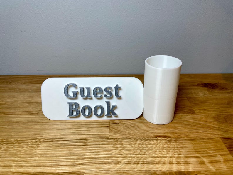 pen pot on the right with a sign that says guest book. Shown on a wooden desk with grey background. White pen pot with a white sign with silver text.