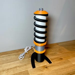 Op de afbeelding: Een zwarte en oranje tafellamp in de vorm van een schokdemper met een witte spiraalveer. De lamp heeft een zwarte voet en een zwart snoer.