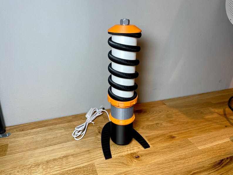 Op de afbeelding: Een zwarte en oranje tafellamp in de vorm van een schokdemper met een witte spiraalveer. De lamp heeft een zwarte voet en een zwart snoer.