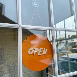 May include: A round orange sign with the word "OPEN" in white letters hanging in a window.