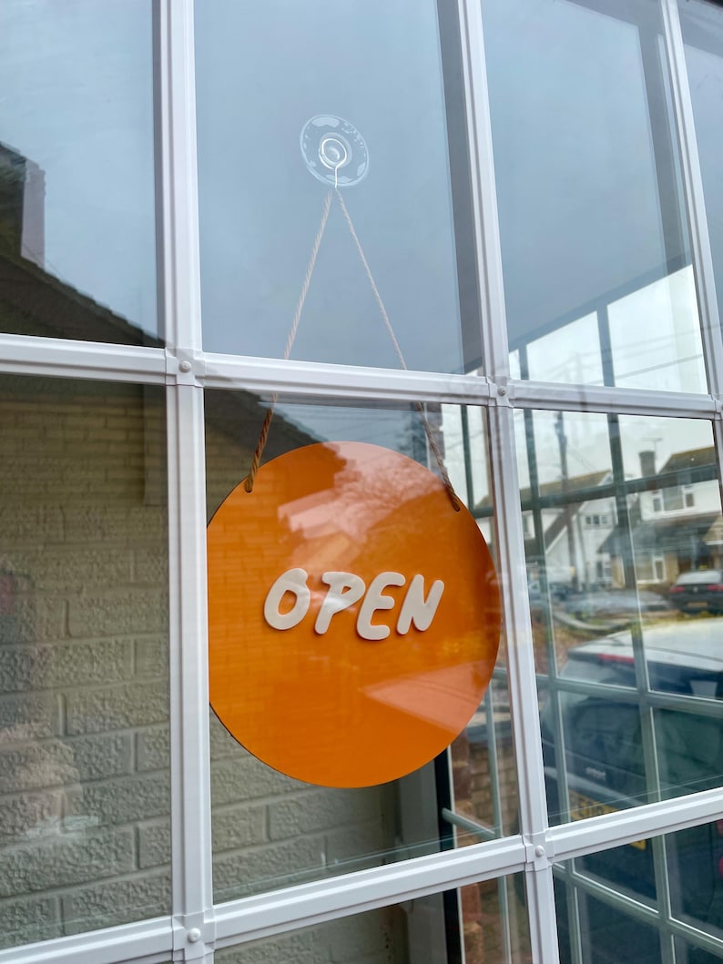 May include: A round orange sign with the word "OPEN" in white letters hanging in a window.