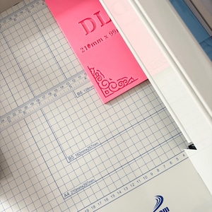 May include: A bright pink rectangular acrylic template with decorative corner flourishes and the text DL 210mm x 99mm, positioned on a white paper cutter with a grid surface featuring measurements in inches and centimeters and the HFS logo.