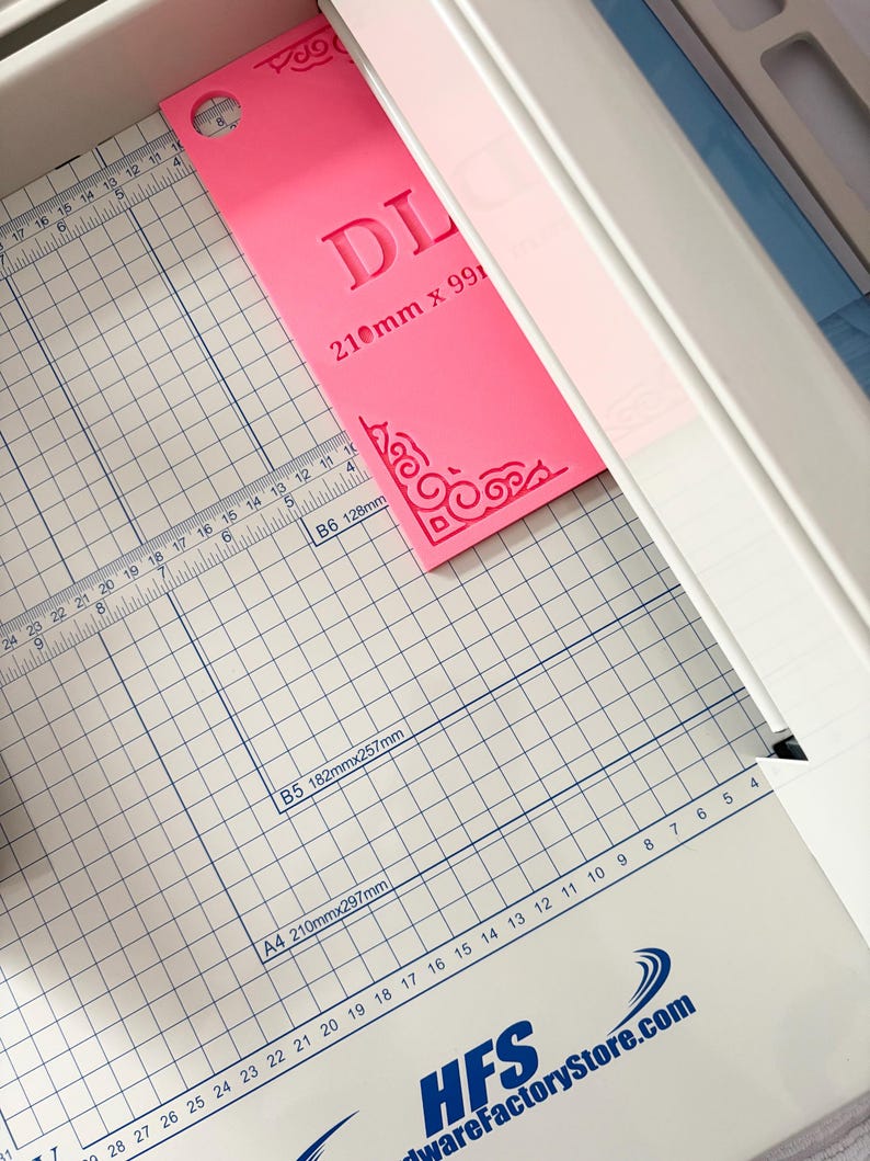 May include: A bright pink rectangular acrylic template with decorative corner flourishes and the text DL 210mm x 99mm, positioned on a white paper cutter with a grid surface featuring measurements in inches and centimeters and the HFS logo.