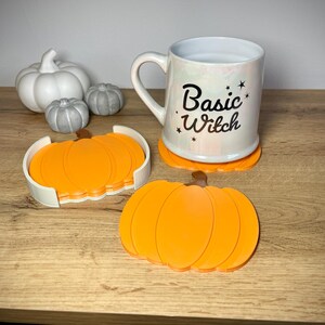Puede incluir: Una taza blanca con el texto "Basic Witch" y acentos de estrellas se encuentra sobre un posavasos de calabaza naranja. Tambi&eacute;n hay posavasos de calabaza naranja adicionales y calabazas decorativas blancas y grises.