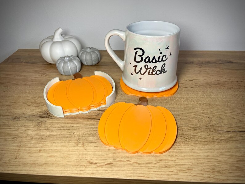 Puede incluir: Una taza blanca con el texto "Basic Witch" y acentos de estrellas se encuentra sobre un posavasos de calabaza naranja. Tambi&eacute;n hay posavasos de calabaza naranja adicionales y calabazas decorativas blancas y grises.
