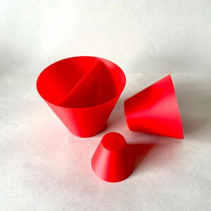 May include: Bright red 3D-printed objects, including a large cone divided into two sections, a smaller cone, and a truncated cone. The objects are arranged on a white surface, showcasing their geometric shapes and vibrant color.