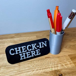 pen pot on the right with a sign that says check in here. Shown on a wooden desk with grey background. Silver pen pot with a black sign with silver text.