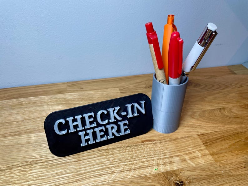 pen pot on the right with a sign that says check in here. Shown on a wooden desk with grey background. Silver pen pot with a black sign with silver text.