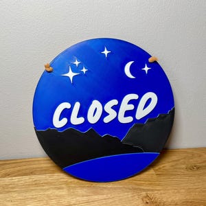 May include: A blue and black round sign with the word "CLOSED" in white lettering. The sign has a crescent moon and stars on a blue background.