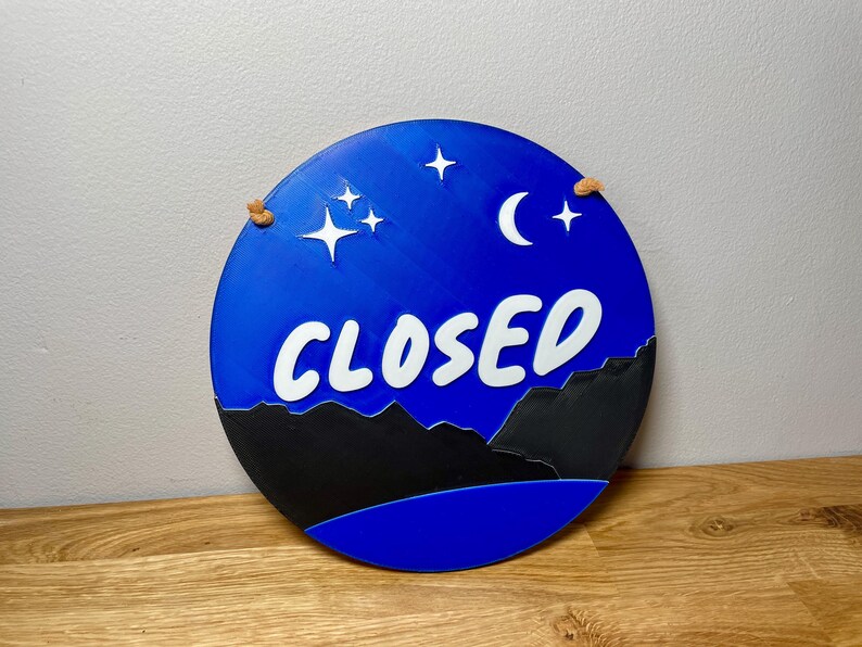 May include: A blue and black round sign with the word "CLOSED" in white lettering. The sign has a crescent moon and stars on a blue background.
