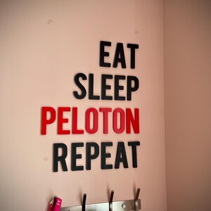May include: Wall decor with the words "EAT SLEEP PELOTON REPEAT" in black and red letters. The letters are affixed to a light pink wall. Below the text are hooks for hanging items.