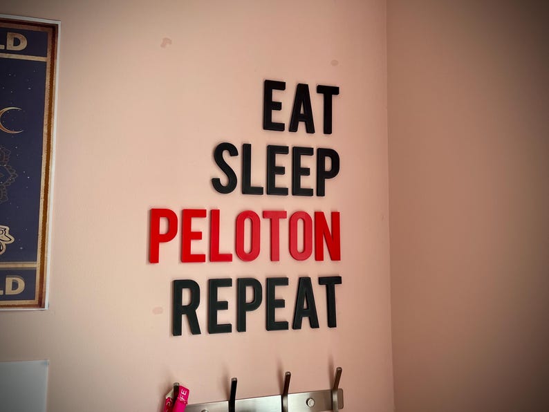 May include: Wall decor with the words "EAT SLEEP PELOTON REPEAT" in black and red letters. The letters are affixed to a light pink wall. Below the text are hooks for hanging items.