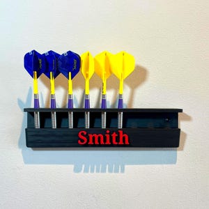 May include: A black dart holder with the name "Smith" in red lettering. The holder contains six darts, three with blue and three with yellow flights. The darts are arranged in a row.