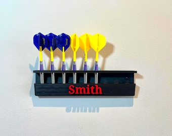 May include: A black dart holder with the name "Smith" in red lettering. The holder contains six darts, three with blue and three with yellow flights. The darts are arranged in a row.