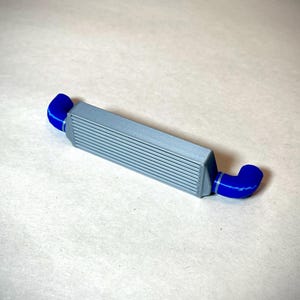 May include: A gray intercooler with blue hoses. The intercooler has a rectangular shape with horizontal lines across the front. The blue hoses are curved and connect to each end of the intercooler. Automotive part.