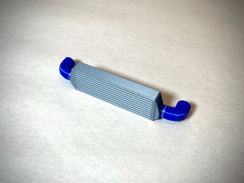May include: A gray intercooler with blue hoses. The intercooler has a rectangular shape with horizontal lines across the front. The blue hoses are curved and connect to each end of the intercooler. Automotive part.