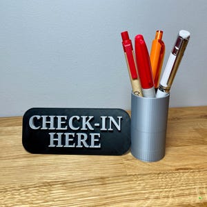 May include: A black sign with the text "CHECK-IN HERE" in white letters. A gray pen holder with four pens inside. The pens are red, orange, white, and brown.