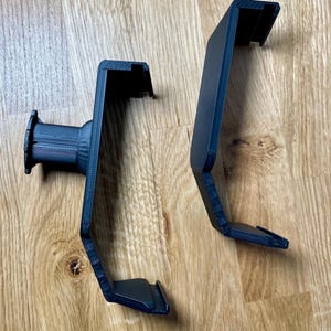 May include: Two black 3D printed phone holders with a textured surface. The holders are designed to be mounted on a wall or other surface.