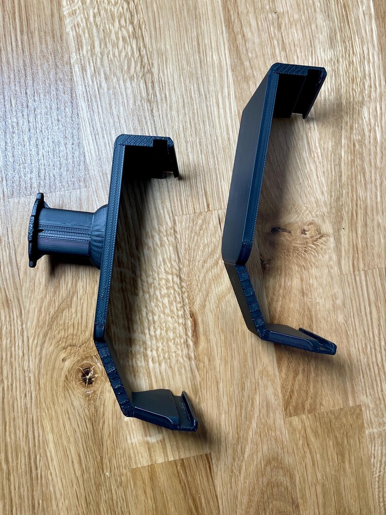 May include: Two black 3D printed phone holders with a textured surface. The holders are designed to be mounted on a wall or other surface.