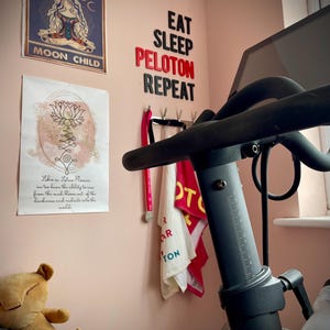 May include: A home gym setup with a Peloton exercise bike. The wall features motivational posters and the words "EAT SLEEP PELOTON REPEAT" in black and red letters. A plush toy and a red Peloton ball are also visible.