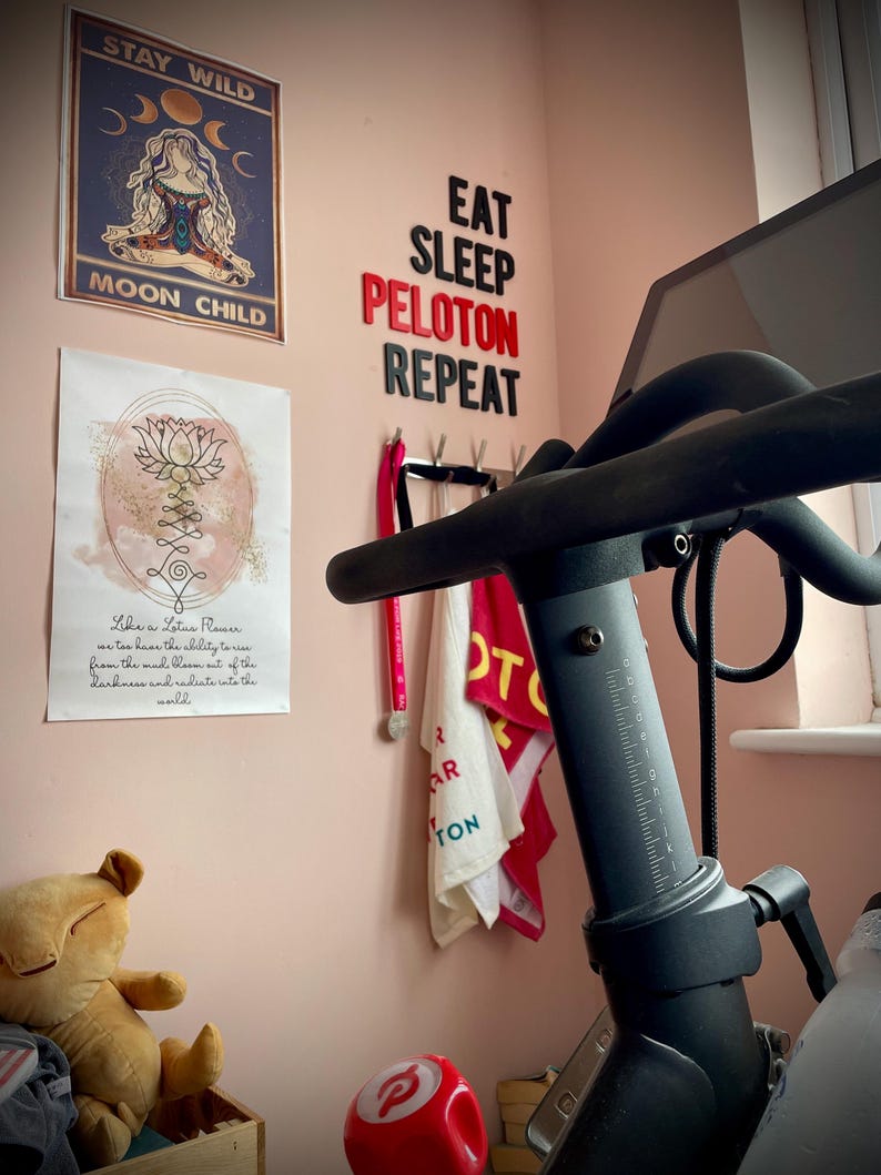 May include: A home gym setup with a Peloton exercise bike. The wall features motivational posters and the words "EAT SLEEP PELOTON REPEAT" in black and red letters. A plush toy and a red Peloton ball are also visible.