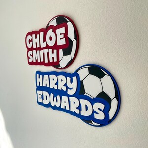 May include: Two personalized soccer-themed wall decorations. One is red with "CHLOE SMITH" in white, and the other is blue with "HARRY EDWARDS" in white. Both have a soccer ball design.