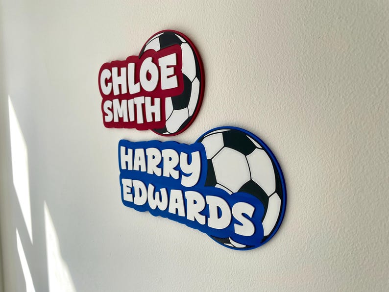 May include: Two personalized soccer-themed wall decorations. One is red with "CHLOE SMITH" in white, and the other is blue with "HARRY EDWARDS" in white. Both have a soccer ball design.